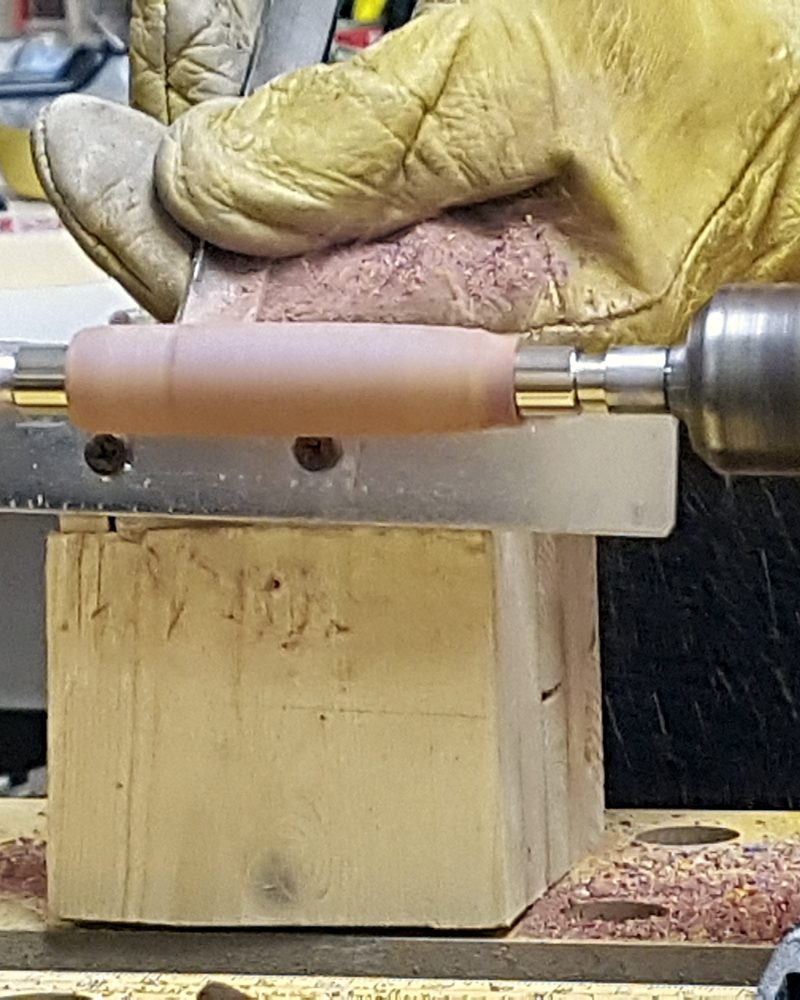 Una mano con il guanto da lavoro rifinisce il fusto di una penna che gira sul tornio